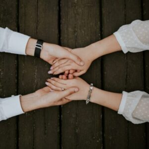 Two people holding hands across a wooden surface. One person wears a watch; the other has a bracelet. Their hands are gently clasped, suggesting comfort or support.