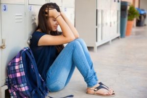Teen girl sits on the floor by lockers, head in hands, looking stressed. A plaid backpack is next to her. She wears jeans, a dark shirt, and sandals.