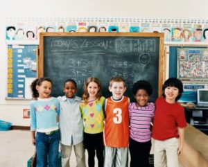 Six young children stand smiling in front of a chalkboard with math problems written on it in a classroom. The children have their arms around each other and appear happy and friendly.