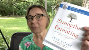 A woman wearing glasses holds up the book Strengths Based Parenting while sitting outdoors, with green grass and trees in the background.