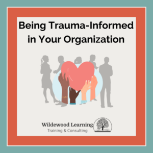 Illustration of diverse hands holding a red heart, with silhouettes of people in the background. Text: Being Trauma-Informed in Your Organization. Wildewood Learning logo at the bottom. Teal border surrounds the image.