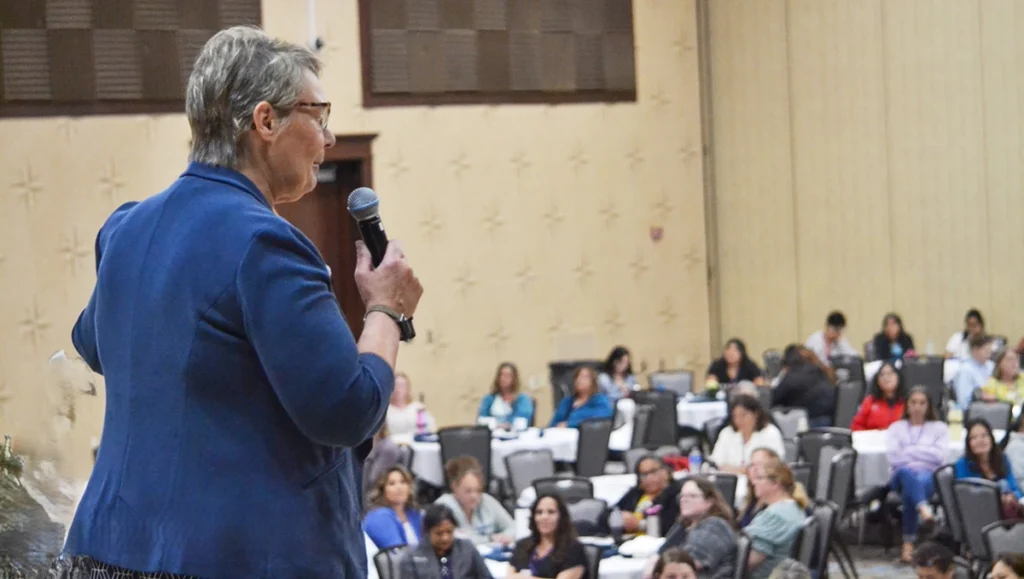 A person with short gray hair speaks into a microphone on stage, addressing an audience seated at round tables in a conference or event hall.