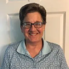 A person with short brown hair and glasses smiles at the camera, wearing a blue patterned collared shirt. They are standing in front of a white paneled door.