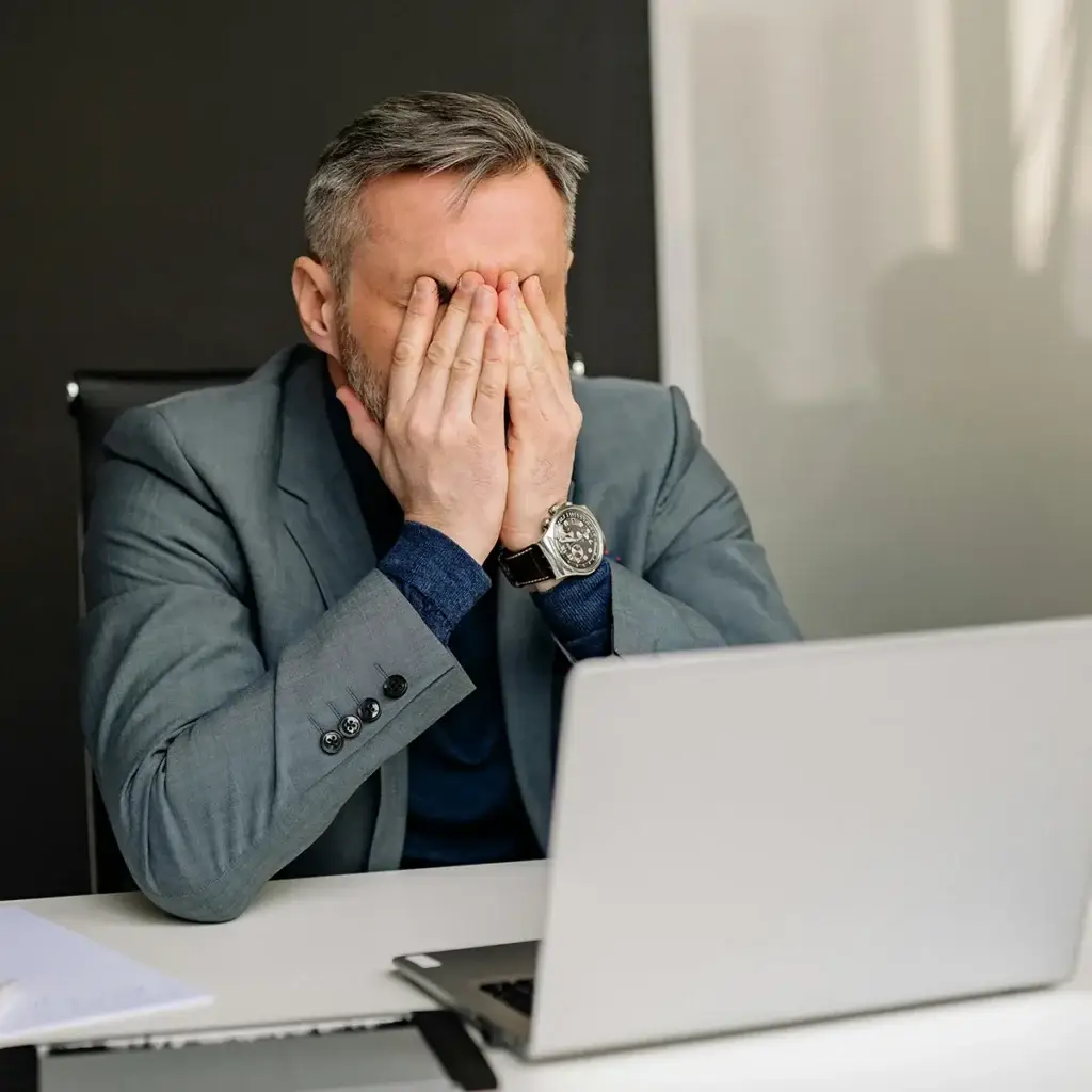 A man in a gray suit sits at a desk with a laptop, covering his face with both hands, appearing stressed or frustrated.