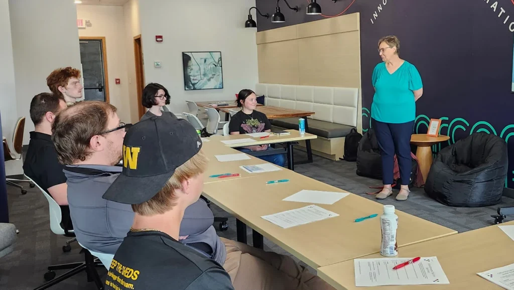 A woman stands speaking to a group of six seated people in a modern meeting room. Papers, pens, and water bottles are on the tables. The atmosphere appears informal and collaborative.