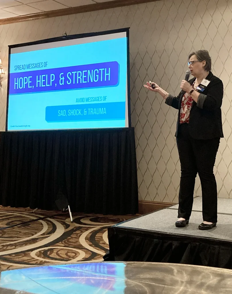 A woman gives a presentation next to a screen displaying the text: Spread messages of hope, help, & strength. Avoid messages of sad, shock, & trauma.