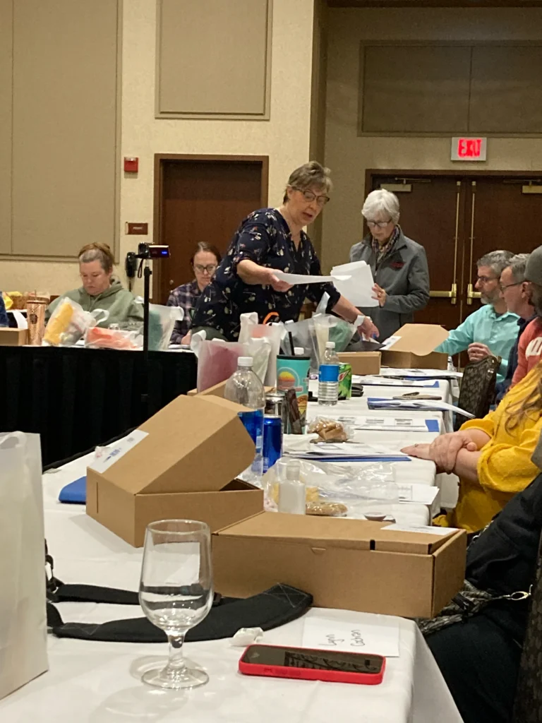 People seated at tables in a conference room with boxes, papers, drinks, and snacks in front of them. Two women stand and look at documents, while others talk or work at the tables. The setting appears to be a workshop or seminar.
