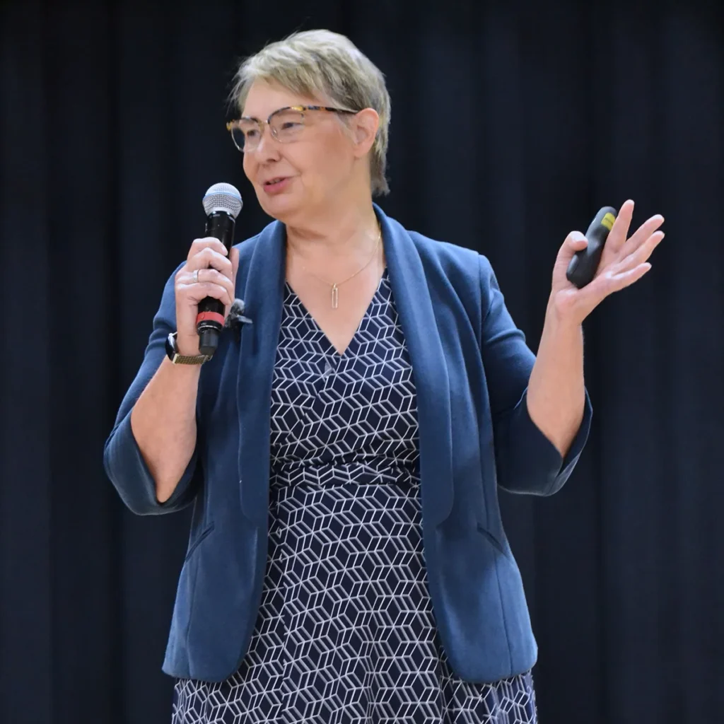 A person with short hair and glasses speaks into a microphone on stage, gesturing with their other hand, against a dark background.