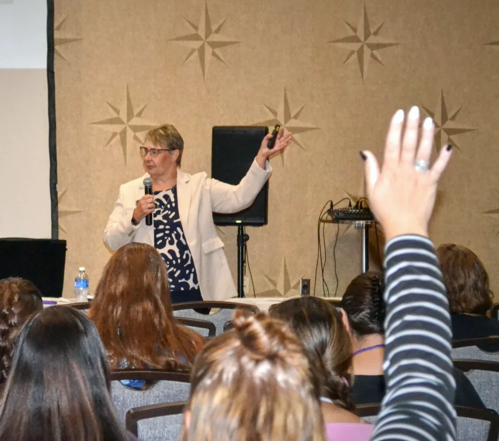 A woman in a light blazer speaks at a conference, pointing forward with a remote. Audience members face her, and one person in the front raises their hand to ask a question.