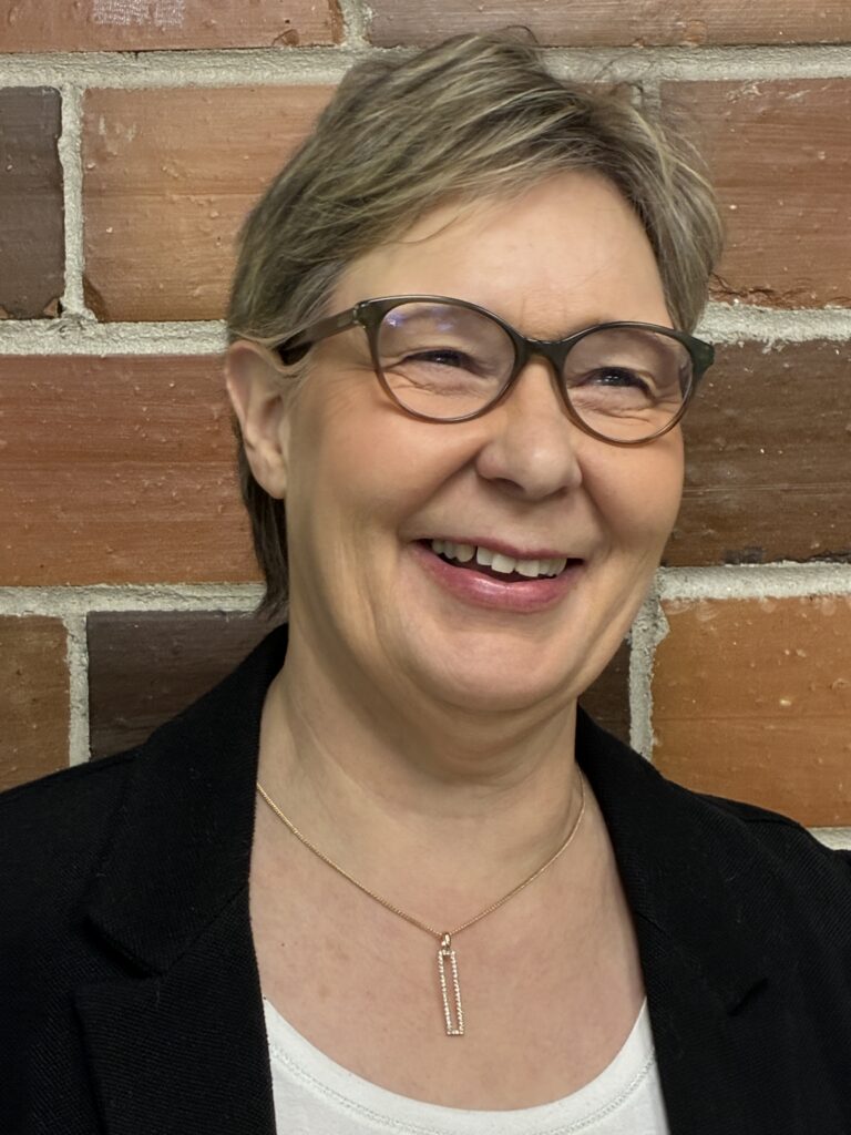 Smiling woman with short, light brown hair and glasses, wearing a black blazer, white top, and gold necklace, standing in front of a brick wall.