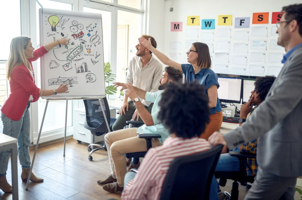 A woman presents ideas on a whiteboard to a group of coworkers in an office. The team looks engaged, some raising hands. The whiteboard has colorful drawings, charts, and the word “TEAM.”.