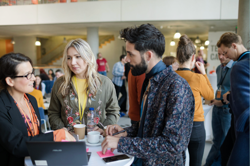 Three people stand around a table with a laptop, coffee cups, and notepads, engaging in conversation at a busy indoor event with others interacting in the background.