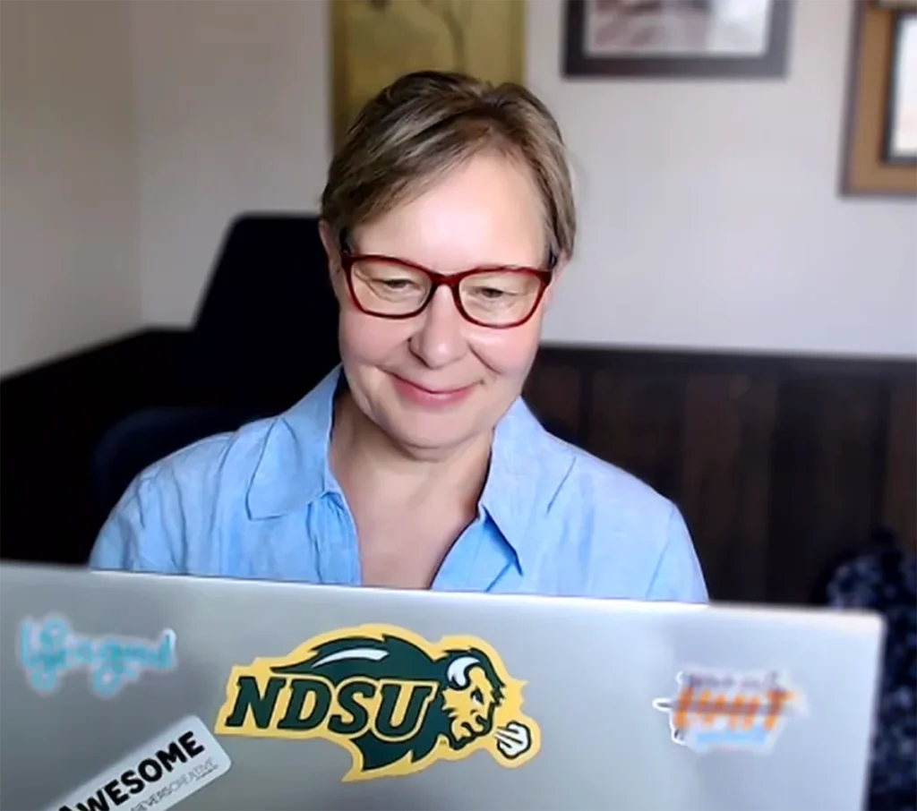 A person with short hair and red glasses smiles while using a laptop covered in stickers, including one that says NDSU. The background is softly blurred, showing a framed picture and dark paneled wall.
